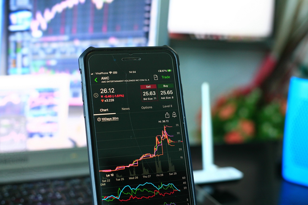 Un smartphone mostrando una interfaz de trading de acciones con gráficos financieros en primer plano y monitores de computadora desenfocados al fondo.